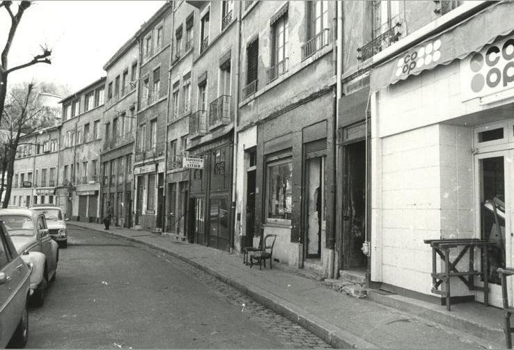 Photographie ancienne en noir et blanc de la devanture : l'enseigne Coop, les voitures des années 1970 garées le long du trottoir